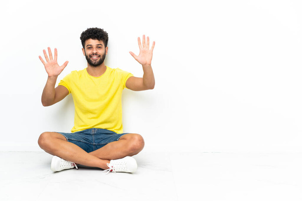 Junger, gut aussehender marokkanischer Mann sitzt auf dem Boden vor isoliertem Hintergrund und zählt mit den Fingern zehn. - Foto, Bild