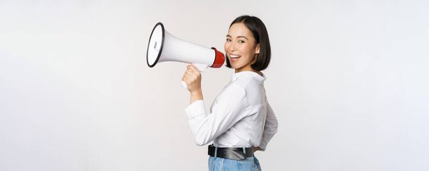 Beautiful young asian woman talking in megaphone, screams in speakerphone and smiling, making announcement, shout out information, standing over white background - Foto, immagini