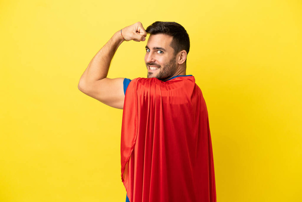 Young handsome caucasian man isolated on yellow background in superhero costume and doing strong gesture - Photo, Image