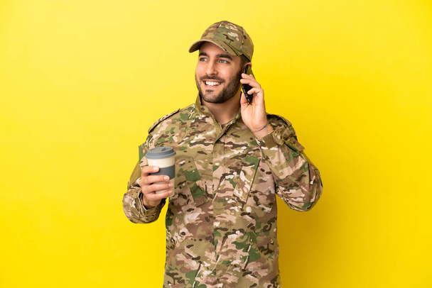 Military man isolated on yellow background holding coffee to take away and a mobile - Photo, Image