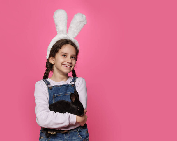 Маленька усміхнена дівчинка в милих великодніх вухах кролика посміхається з щасливим обличчям тримає в руках милий чорний маленький кролик
. - Фото, зображення