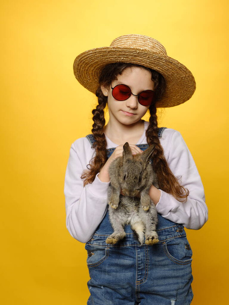 Little girl dressed in a jeans and straw hat and red sunglasses, holding a real rabbit, isolated yellow background. - Фото, зображення