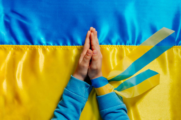 子供たちはウクライナの旗の背景で祈ってリボンで手。平和のシンボルとウクライナのために祈る。トップビュー、フラットレイアウト。手に焦点を当てる. - 写真・画像