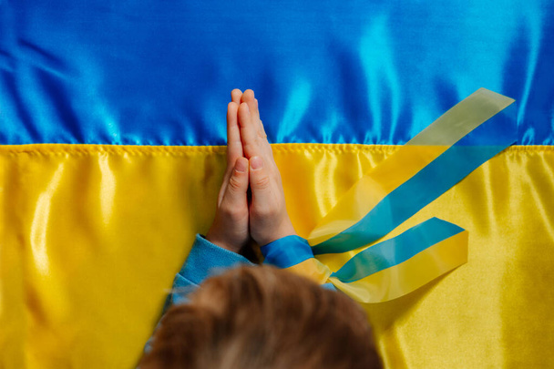 Kids hands with ribbon praying on the Ukrainian flag background. Symbol of peace and pray for Ukraine. Top view, flat lay. Selective focus on the hands. - Photo, Image