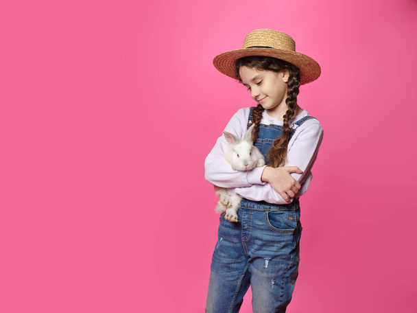 Чарівна кучерява маленька дівчинка в солом'яному капелюсі, тримає справжнього милого кролика, дивлячись на камеру, ізольований рожевий фон
. - Фото, зображення
