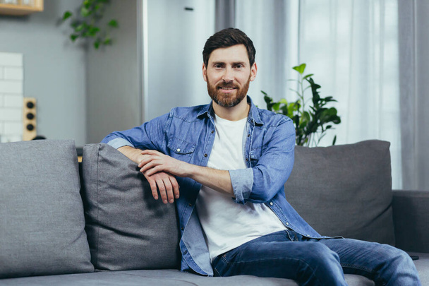 Portrait of a man with a beard at home, sitting on the couch and looking at the camera - Photo, image