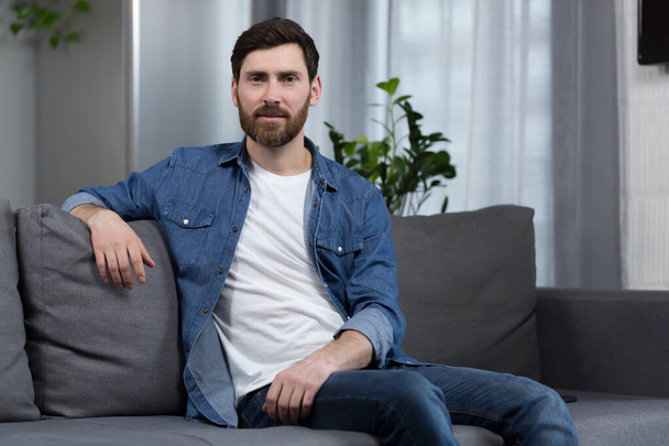Portrait of a man with a beard at home, sitting on the couch and looking at the camera - Foto, Imagen
