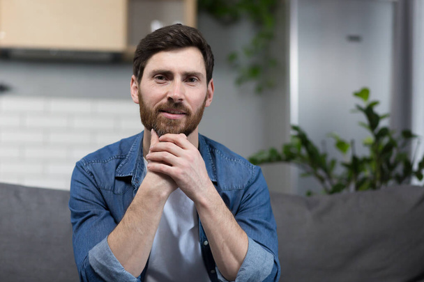 Portrait of a man with a beard at home, sitting on the couch and looking at the camera - Photo, Image