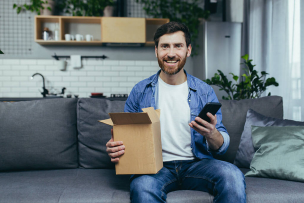 man at home received an order from the online store, happy opens a cardboard box with a parcel, leaves a positive response by phone - Valokuva, kuva