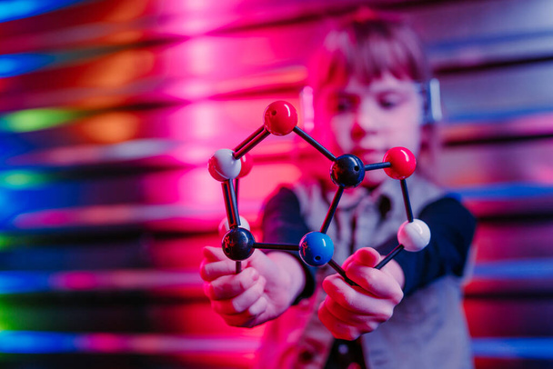 Petite fille tenant modèle moléculaire debout sur le fond coloré néon lumières. Génération Alpha utilisant la technologie. Focus sélectif sur le modèle de boules et bâton en plastique. - Photo, image
