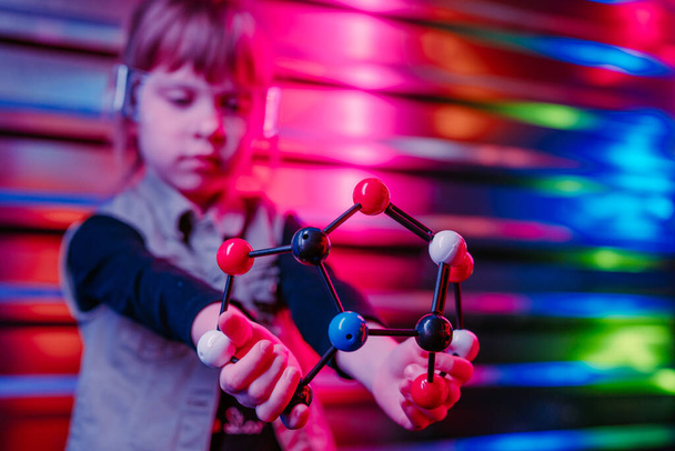 Маленькая девочка с молекулярной моделью, стоящей на фоне ярких неоновых огней. Поколение Альфа с использованием технологии. Селективный акцент на пластиковые шары и модель палки. - Фото, изображение