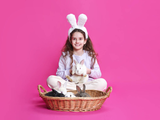 Pełen obraz uśmiechniętej dziewczynki z białymi króliczymi uszami, trzymającej króliczka w rękach, odizolowanej na różowym tle. Przestrzeń kopiowania. - Zdjęcie, obraz
