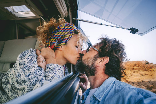 Молодая взрослая пара влюблённая целуется и наслаждается отдыхом в путешествии вместе. Женщина целует мужчину через окно в машине. Туристическая и фургонная жизнь современная альтернативная концепция жизни. Отношения - Фото, изображение