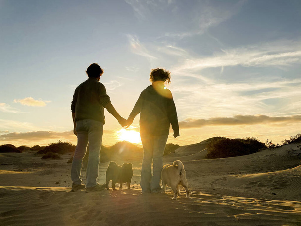 Romantic family with man and woman and two dogs admire and enjoy a colored sunset in outdoor leisure activity. Concept of travel and amazing destination for people. Couple in love together and sun - Photo, Image