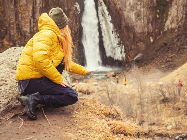 A woman in a yellow jacket squatting on the background of autumn nature put her hand to a waterfall flowing in the rocks in the distance. Soft focus. Optical illusion - Photo, Image
