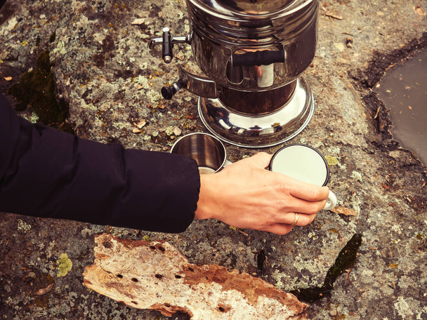 Рука чоловіка розміщує білий залізний кухоль на камені біля блискучого хромованого самовара. Чаювання в природі в осінній день - Фото, зображення