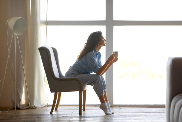 Vista lateral de la hermosa mujer joven bebiendo café caliente, sentado en la silla cerca de la ventana en casa, espacio libre - Foto, Imagen