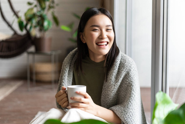 Relajada mujer asiática feliz en ropa acogedora, beber té y sentarse junto a la ventana, mirando hacia el exterior con sonrisa complacida - Foto, imagen