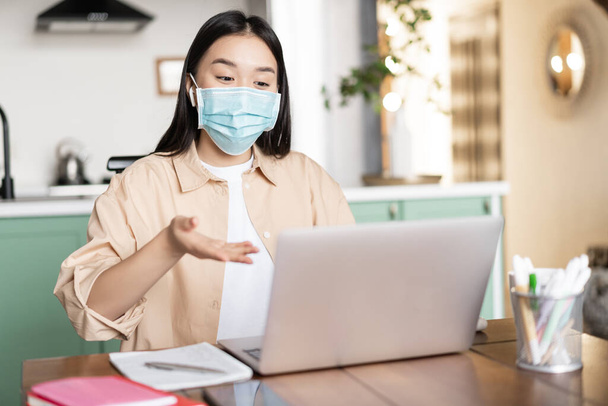 Young asian girl talking at laptop in medical mask, sitting at home, work or study remotely - Valokuva, kuva