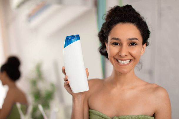Mujer sonriente mostrando botella de champú con maqueta para el diseño en la cámara, loción para el cuidado del cabello publicidad o gel de ducha - Foto, imagen