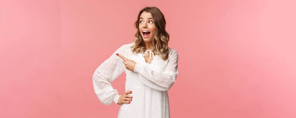 Excited and amazed blond caucasian girl in kawaii dress, open mouth gasping in awe, pointing and looking left with excitement and astonishement, see great beauty product, pink background - Photo, Image