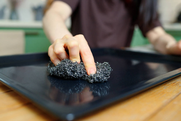 Close-up em uma mão com uma esponja de metal que limpa uma assadeira do forno. - Foto, Imagem