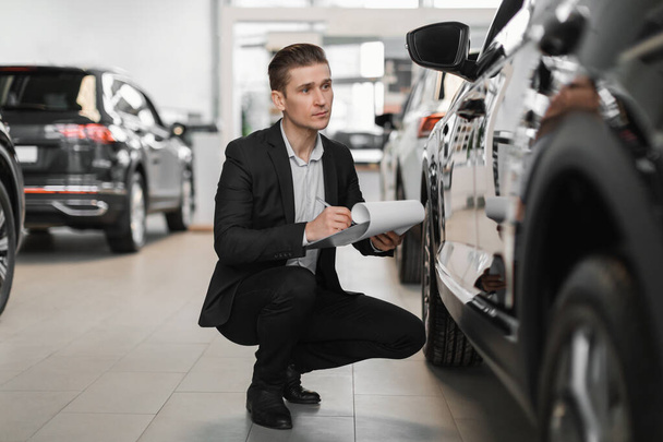 Auto agent ubezpieczeniowy siedzi w pobliżu nowego samochodu, sprawdzając go, robiąc notatki, pracując w centrum sprzedaży samochodów - Zdjęcie, obraz