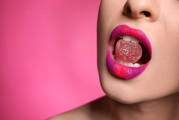 Young woman with beautiful makeup holding candy in lips on pink background, closeup. Space for text - Foto, Bild