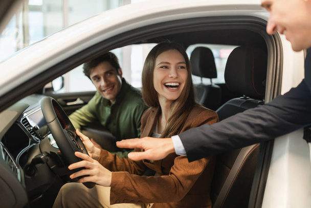 Ευτυχισμένο νέο ζευγάρι δοκιμή οδήγησης νέο αυτοκίνητο, πωλητής βοηθώντας τους να κάνουν την καλύτερη επιλογή στο auto αντιπροσωπείας - Φωτογραφία, εικόνα