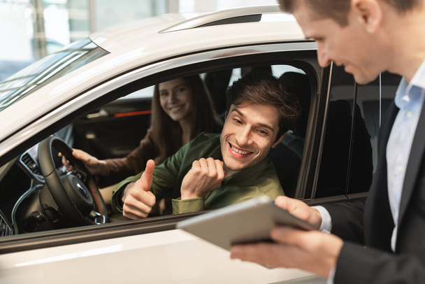 Junges Paar besucht Autosalon, sitzt im Auto, spricht mit Verkäuferin, schaut auf Tablet im Autohaus - Foto, Bild