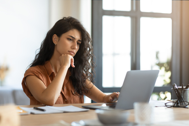 Bedachtzame jonge brunette vrouw op zoek naar laptop scherm, werken online op kantoor in huis kantoor, vrije ruimte - Foto, afbeelding