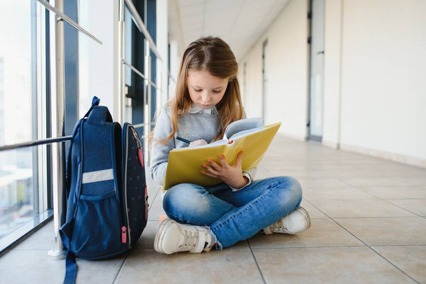 Vue de face de jolie écolière blonde tenant beaucoup de notes et de livres colorés. Adolescente intelligente souriant à la caméra, debout sur le couloir de l'école internationale. - Photo, image