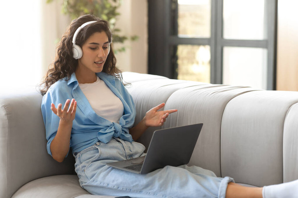 Pretty millennial woman in headphones communicating online on laptop, having video conference indoors, free space - Photo, Image