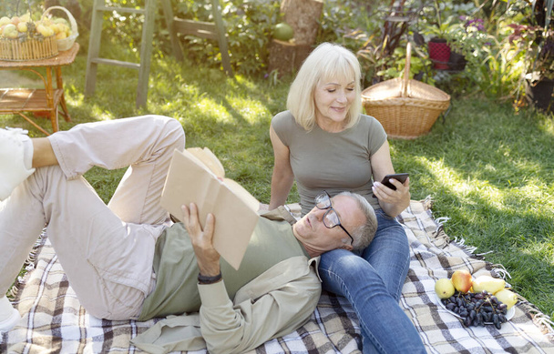 Retired spouses resting in their garden, woman using smartphone while man reading book, lying on wifes laps - Фото, изображение