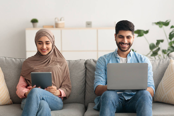 Μουσουλμανικό ζευγάρι που περιηγείται στο διαδίκτυο στο φορητό υπολογιστή και ψηφιακή ταμπλέτα εσωτερικά - Φωτογραφία, εικόνα