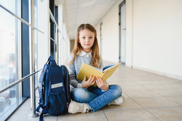 Front view of pretty blonde school girl holding many colorful notes and books. Clever teen girl smiling at camera, standing on corridor of international school. - Zdjęcie, obraz