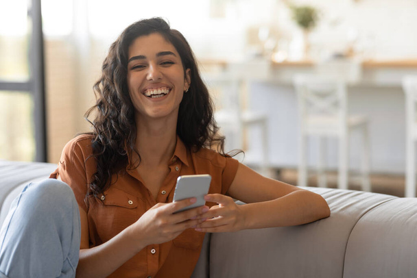 Gelukkige jonge vrouw zitten op de bank in de woonkamer, met behulp van smartphone, chatten met een vriend, communiceren online - Foto, afbeelding