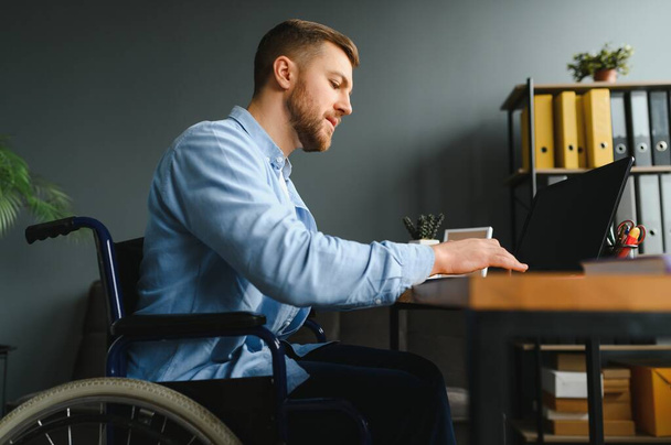 Disabled person in the wheelchair works in the office at the computer. He is smiling and passionate about the workflow. - 写真・画像