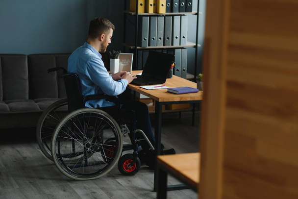 Young man with special needs in casual clothes working on wireless laptop. Male freelancer working from home while sitting in wheelchair. - Foto, afbeelding