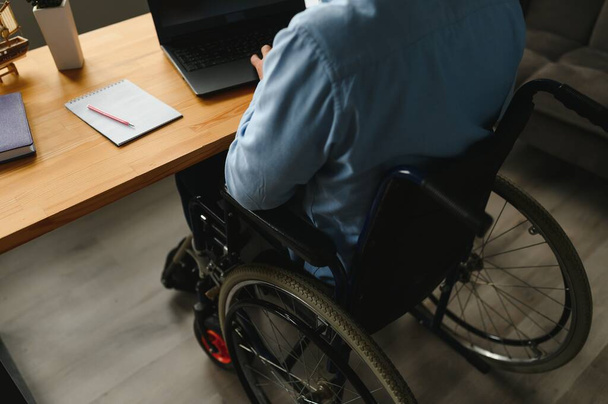 Young man with special needs in casual clothes working on wireless laptop. Male freelancer working from home while sitting in wheelchair. - Foto, imagen