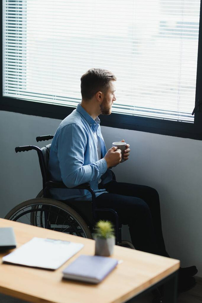 Disabled Person Sits in Wheelchair Against Window. Serious Sad Caucasian Man Wearing Casual Clothes and Look at Large Panoramic View in Bright Modern Living Room or Hospital. - Photo, Image