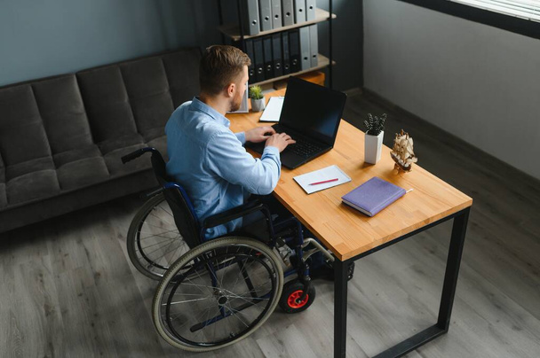 Young man with special needs in casual clothes working on wireless laptop. Male freelancer working from home while sitting in wheelchair. - Foto, immagini