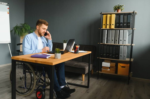 Disabled person in the wheelchair works in the office at the computer. He is smiling and passionate about the workflow. - Foto, afbeelding