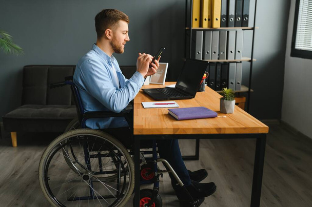 Disabled person in the wheelchair works in the office at the computer. He is smiling and passionate about the workflow. - Foto, Imagem