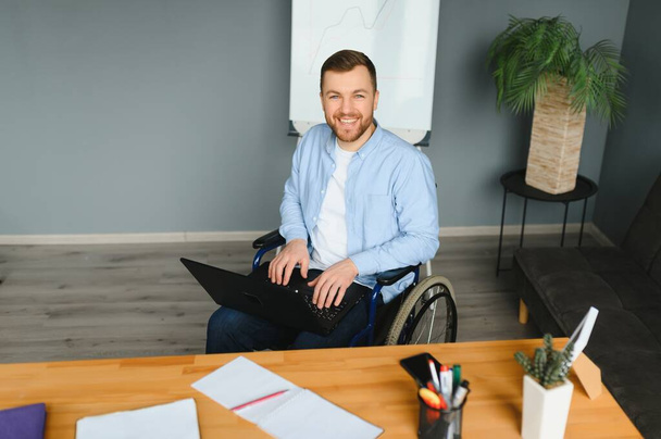 Disabled person in the wheelchair works in the office at the computer. He is smiling and passionate about the workflow. - Photo, image