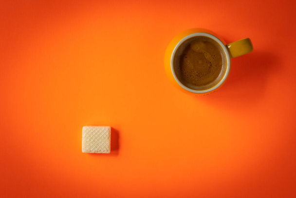 Taza amarilla de café espresso doble recién hecho sobre fondo naranja con oblea de tamaño partido en el lado opuesto del marco - Foto, Imagen
