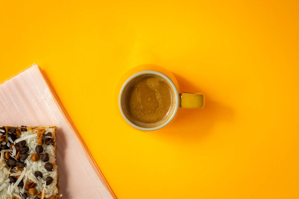 Yellow double espresso coffee cup in the center of bright yellow background. Layered baked pastry decorated with coconut pills and chocolate chips placed on backing sheet in the corner. - Фото, изображение