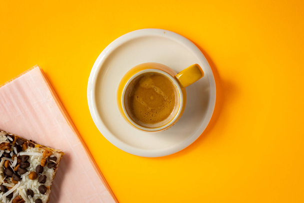 Een mok van vers gebrouwen dubbele espresso geplaatst op een schotel in het midden van platte heldere gele achtergrond. Gelaagde taart versierd met kokospillen en chocolade chip geplaatst op bakpapier. - Foto, afbeelding