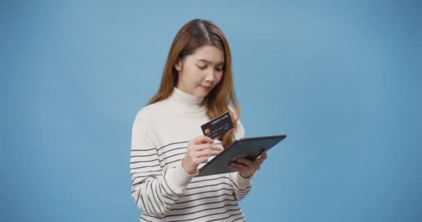 Nuoret iloinen Aasian nainen käyttää rento villapaita internet ostoksia nainen verkossa digitaalinen tabletti ja luottokortti seistä eristetty yli sininen tausta. Naisten käyttö pankkikorttimaksu verkkopankki. - Materiaali, video