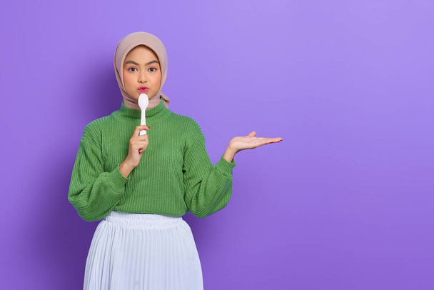 Fröhlich schöne asiatische Frau in grünem Pullover und Hijab hält einen Löffel und zeigt Kopierraum auf Händen isoliert über lila Hintergrund - Foto, Bild